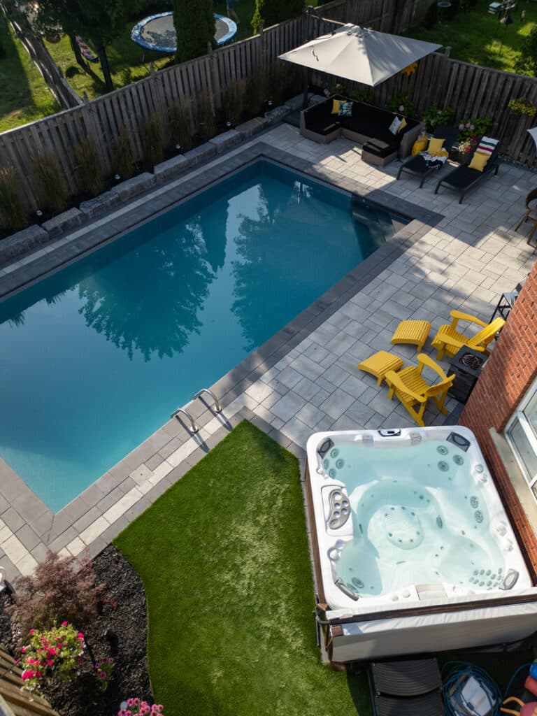 A backyard landscape build by OGS, it features a swimming pool, tilework, a hot tub, stonework, garden, grass area, chairs and an umbrella.
