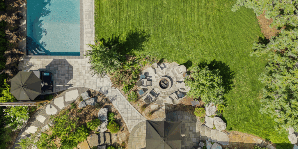 An overhead aerial view of a luxury backyard featuring a geometric swimming pool and a circular stone fire pit area. The pool is bordered by light grey square pavers. A winding walkway made of smaller rectangular pavers connects the pool area to a circular flagstone patio surrounding a central metal fire pit. Large natural boulders are strategically placed among lush green garden beds to provide structural interest. Two large black patio umbrellas provide shade over seating areas, and the entire space is bordered by a perfectly manicured vibrant green lawn.