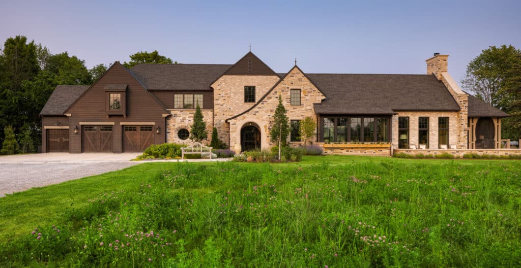 A wide front view of a large stone and dark wood estate featuring a diverse landscape. In the foreground, a lush, naturalized meadow with green grass and purple clover transitions into a manicured lawn. The home's entrance is highlighted by a formal stone archway, flanked by symmetrical upright trees and curated garden beds. A wooden garden bench sits on a flagstone landing near the driveway, which transitions from grey gravel to smooth concrete. The plantings include a mix of ornamental grasses, lavender, and perennial shrubs that soften the transition between the stone facade and the lawn.