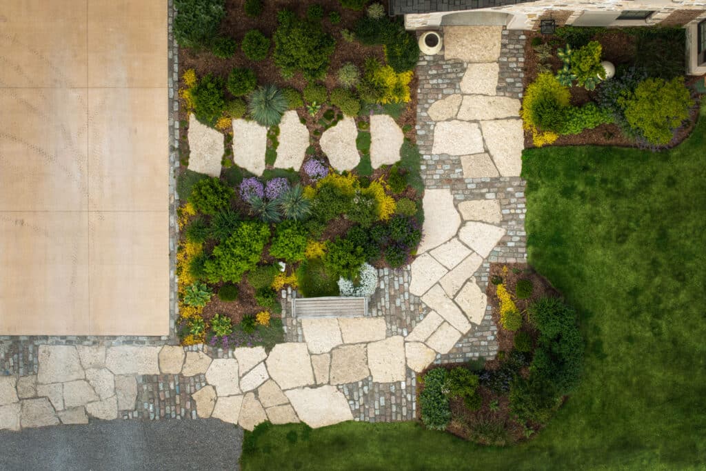 A high-angle vertical shot of a custom-designed walkway and garden bed. The path is constructed using large, irregular cream-colored flagstones inlaid with sections of small grey cobblestones, creating a textured, "old-world" mosaic effect. The walkway leads to a stone arch entrance. Adjacent to the path is a dense garden bed filled with a variety of textures: purple creeping phlox, lime-green groundcovers, dark green mounded shrubs, and silvery-blue succulents, all set against a dark organic mulch. A weathered wooden bench is tucked into a dedicated flagstone nook within the garden.