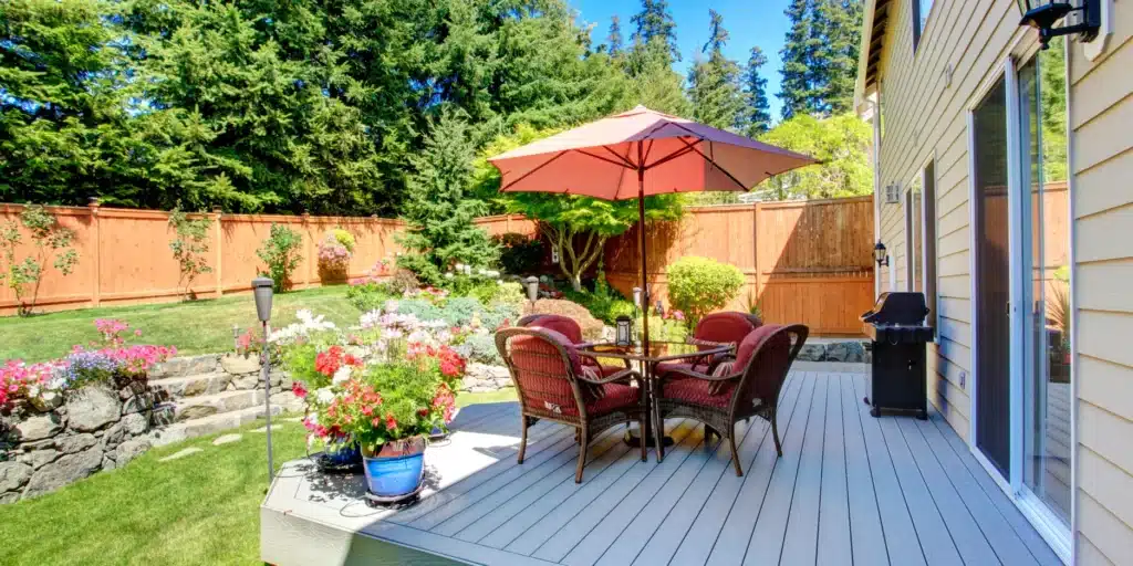A spacious, elevated composite deck designed by OGS Landscapes for outdoor dining, featuring integrated planters, a privacy fence, and tiered stone retaining walls with blooming summer flowers.