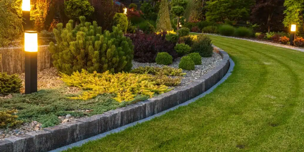 A professional OGS Landscapes garden bed featuring architectural evergreens and gold-toned shrubs, accented by modern bollard lighting and a crisp stone edge against a perfectly striped lawn.