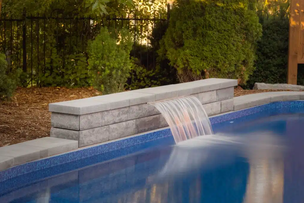 A focused shot of an integrated sheet waterfall feature made of grey stacked stone overflowing into a blue-lined pool, illustrating the durable, high-quality material selection and professional pool construction services essential for timeless landscapes.
