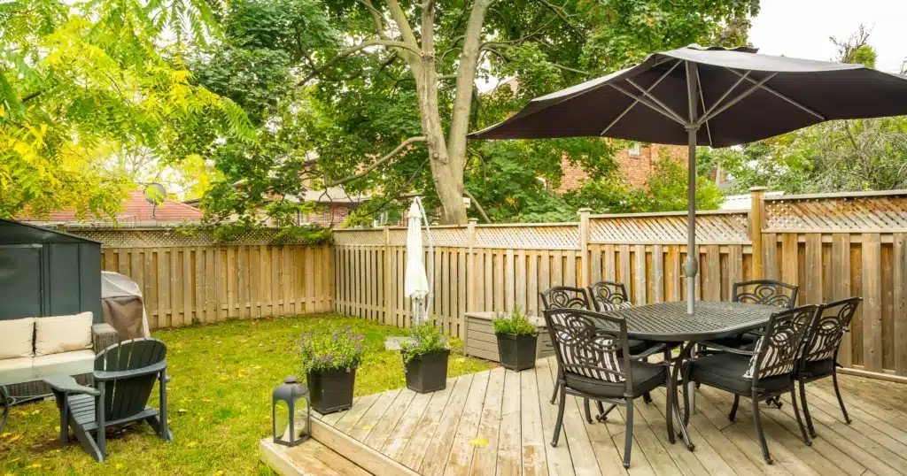 OGS Landscape created this cozy wooden deck and patio combo. The design features a dark metal dining set under a large umbrella, comfortable rattan lounge seating on the grass, a charcoal BBQ, and potted plants, all enclosed by a private wooden lattice fence.