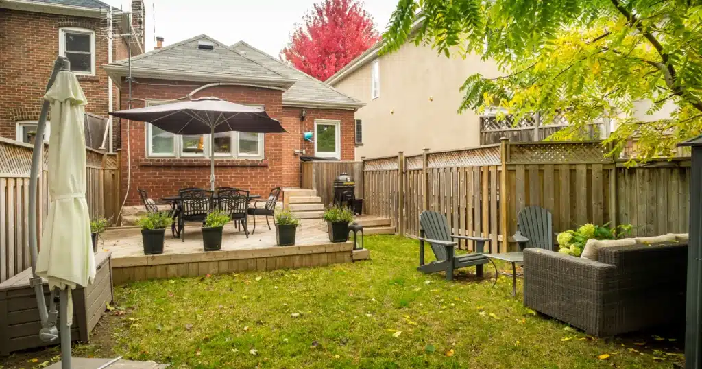 A tiered outdoor entertaining area by OGS Landscape in a suburban backyard. A multi-level wood deck supports a dining table with an umbrella and several potted plants. The lower grass level includes grey Adirondack chairs and a modern rattan sofa, flanked by private fencing and seasonal red foliage.