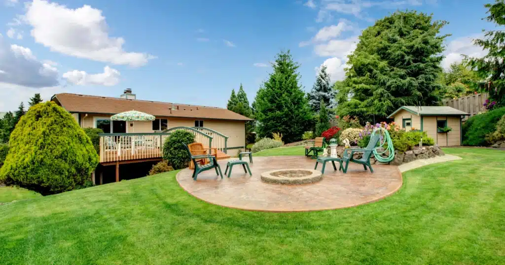 A expansive backyard designed by OGS Landscape, highlighting a circular stamped concrete patio with an integrated stone fire pit and surrounding lounge chairs. The space is set on a lush green lawn next to a custom wood deck with a pergola, all integrated into a hillside landscape.